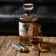 (product) English Pewter Company x He Spoke Style 2 oz. Flask displayed with a bottle of whisky and a glass on a rustic wooden surface
