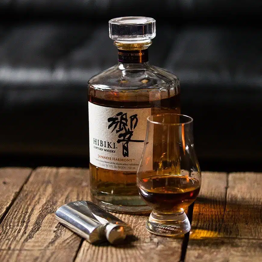 (product) English Pewter Company x He Spoke Style 2 oz. Flask displayed with a bottle of whisky and a glass on a rustic wooden surface