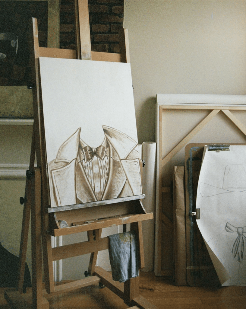 Easel featuring a detailed sketch of a suit jacket and shirt Easel featuring a detailed sketch of a suit jacket and shirt, with additional art materials and canvases leaning against a wall in a studio setting
