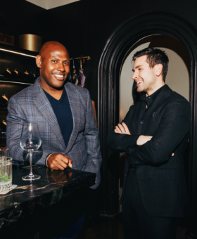 Two men dressed in tailored eveningwear—one in a navy check sport coat, the other in an all-black suit—enjoying drinks at Michael Andrews Bespoke during “A Night in Portofino” event