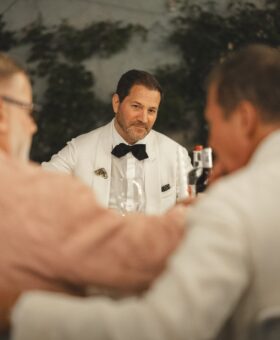 Guests in tailored eveningwear seated around a candlelit dinner table, with Michael in a white tuxedo jacket and black bow tie at the Pitti SS 2024 Arno Dinner