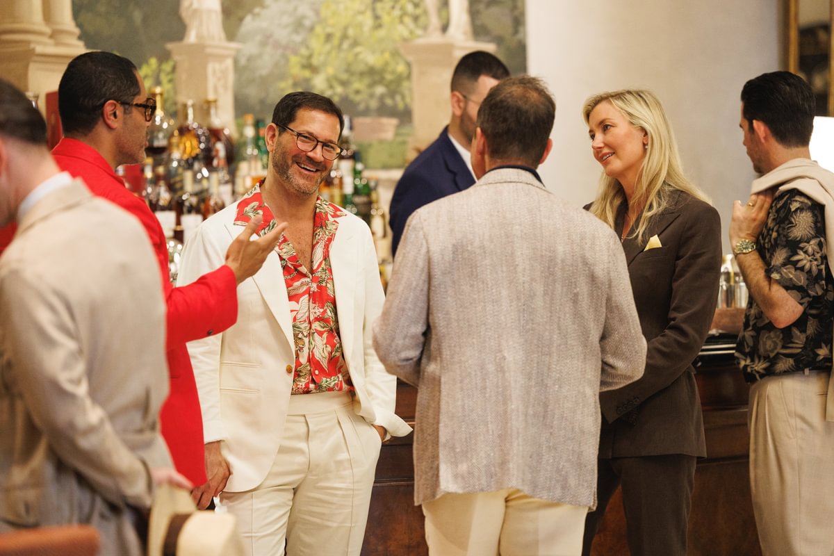 Group of well-dressed guests mingling at a bar during Pitti Uomo, featuring Michael in an ivory suit with a red tropical shirt