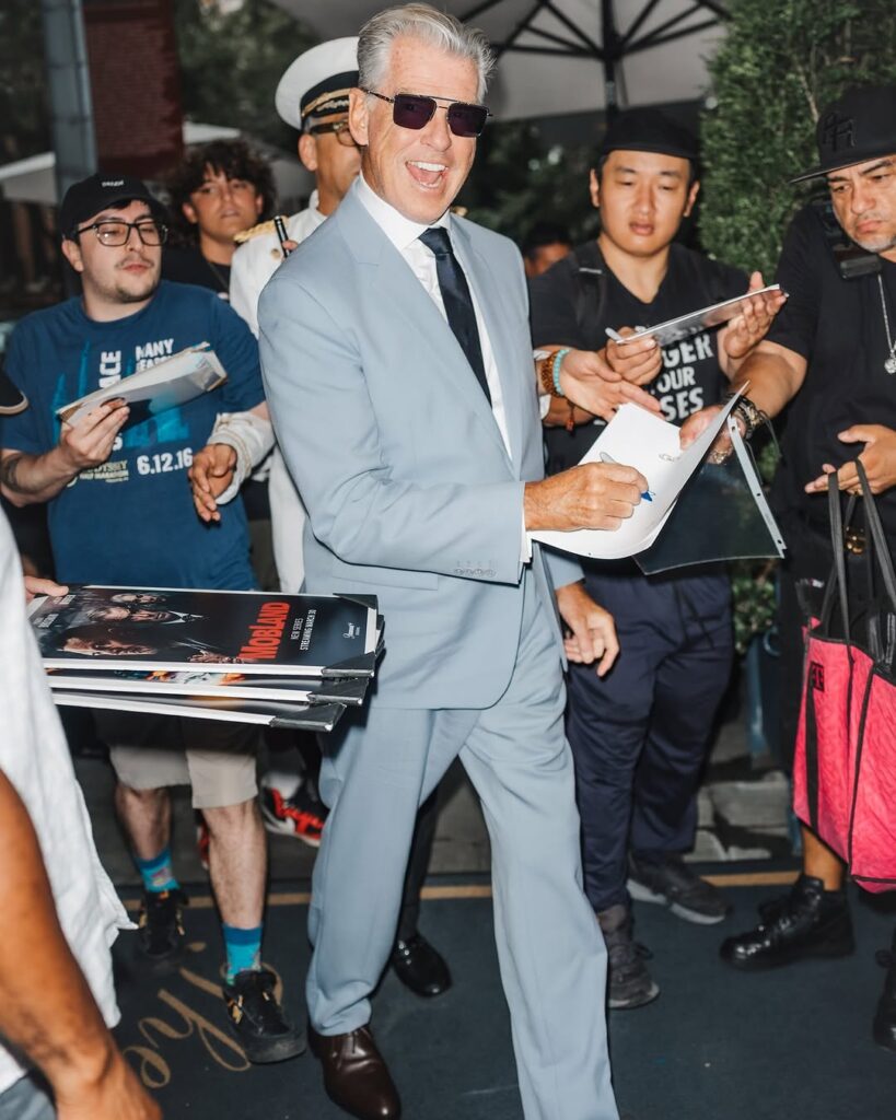 Pierce Brosnan smiling in a blue suit while signing autographs—tailored, charismatic, and Bond-ready in New York City.