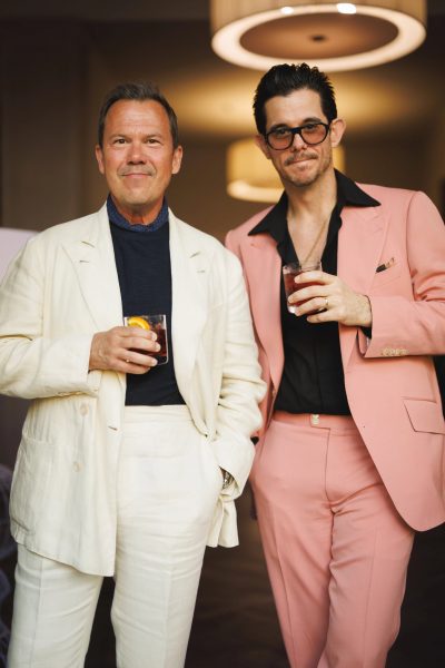Two men in tailored suits holding cocktails at an indoor event; one wears a cream suit with a navy shirt, the other a pink suit with a black shirt and sunglasses. - custom suits nyc bespoke tailor midtown, noho