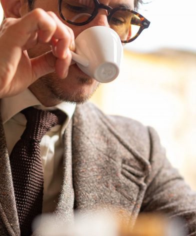 Brown Donegal tweed suit with flecked weave, cream shirt and brown knit tie, worn by a model sipping espresso