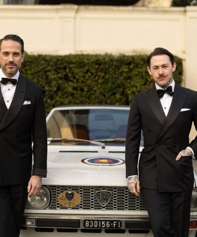 Black double-breasted tuxedos with shawl and peak lapels and bow ties, worn by two models by a vintage car