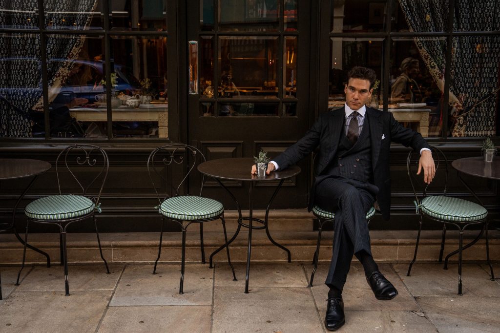 MAB ambassador Austin Pollak seated outside a London café in a midnight green herringbone three-piece suit crafted from Dormeuil Amadeus cloth.