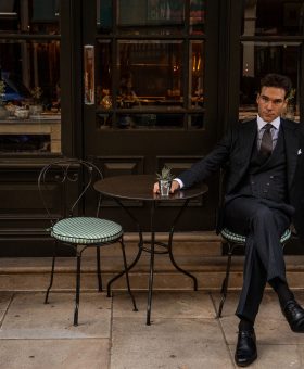 MAB ambassador Austin Pollak seated outside a London café in a midnight green herringbone three-piece suit crafted from Dormeuil Amadeus cloth.