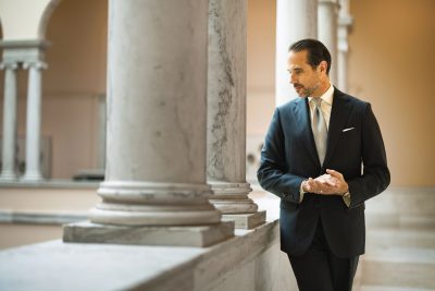 Brian Sacawa wearing a tailored navy suit in a marble-columned interior for Michael Andrews Bespoke’s 20 for 20 Look Book.
